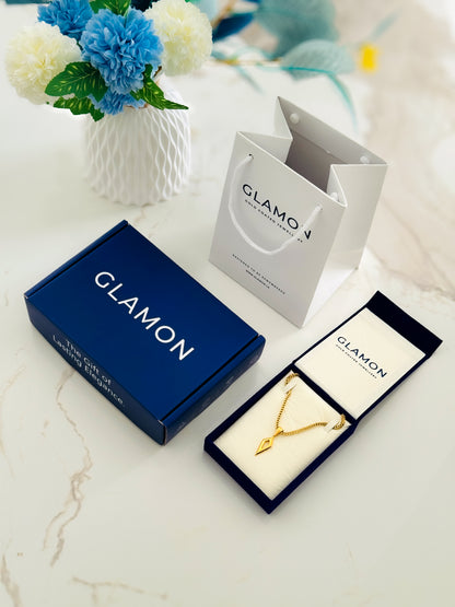 A gold necklace with a geometric pendant displayed in an open navy jewelry box, alongside a navy brand box, a white branded shopping bag, and a vase of blue and white flowers on a light marble surface.
